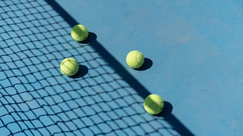 Four yellow tennis balls resting on a bright blue hard court in sunshine, with the shadow of the net falling diagonally across the surface