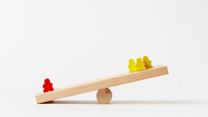 A plank of pale wood balanced on a round wooden block, tipped down at the left end where one red peg figure stands alone, while three yellow peg figures stand together at the raised right end, against a plain white background