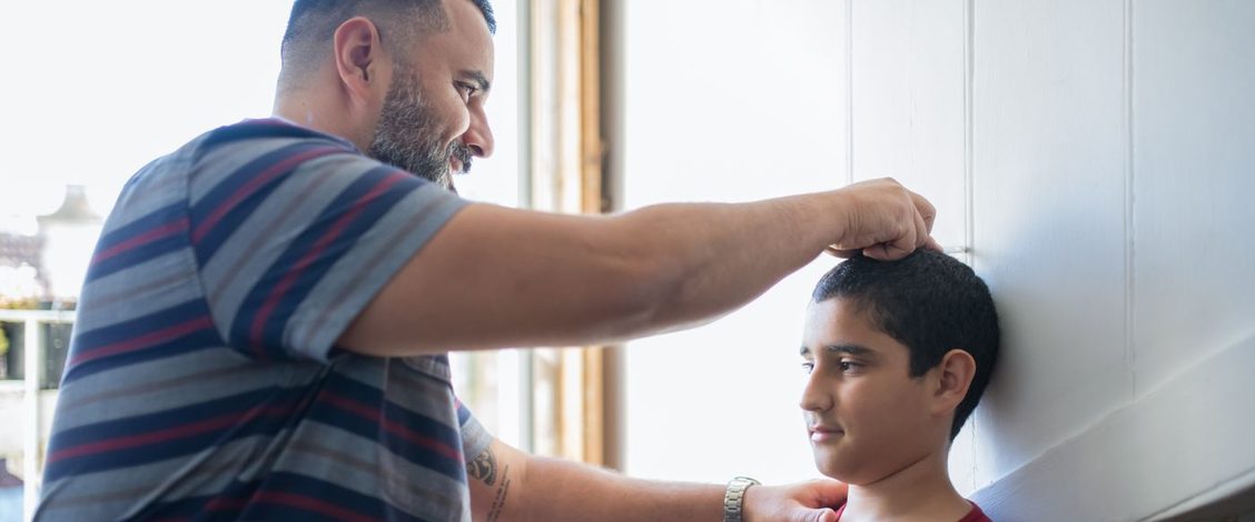A father in a striped T-shirt marks his son&rsquo;s height against a white wall in a sunlit room while the boy stands straight with his back to the wall