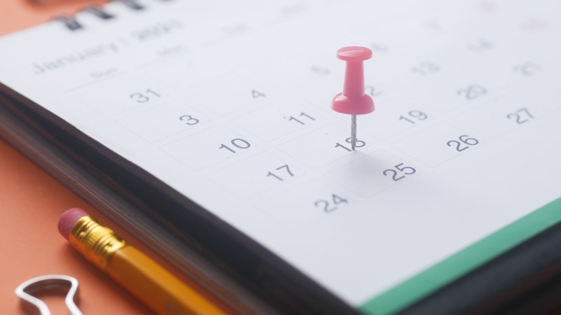 A pink push pin stuck into one date on a calendar page, with a yellow pencil and two paper clips on an orange background.