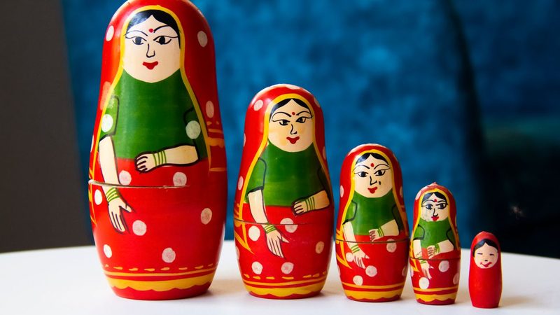 Five red and green painted nesting dolls of the same woman standing in a row from largest to smallest