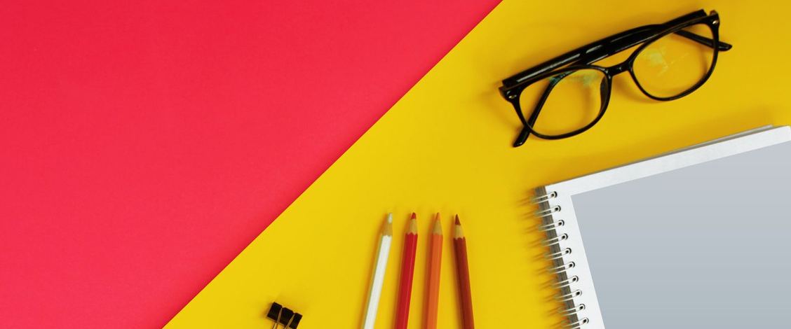 Black framed reading glasses set down on a yellow and red surface beside four coloured pencils, two binder clips and a spiral notebook
