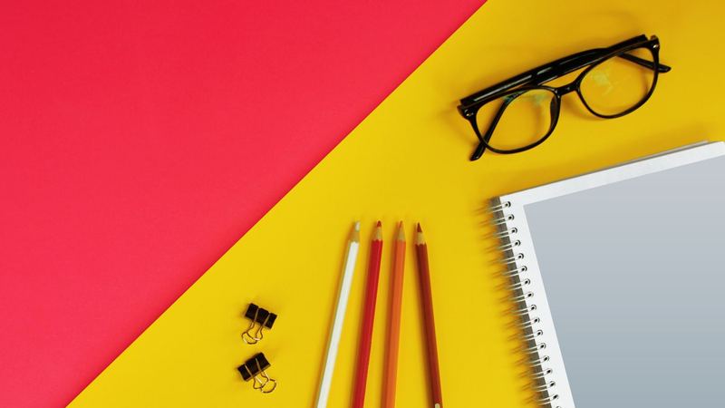 Black framed reading glasses set down on a yellow and red surface beside four coloured pencils, two binder clips and a spiral notebook