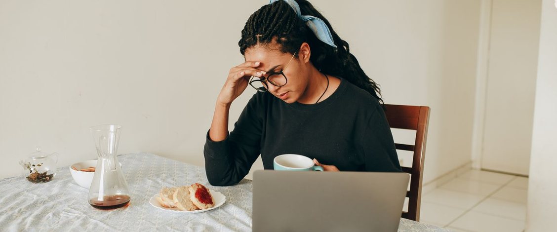 A woman with braided hair tied back with a pale blue scarf sits at a kitchen table in daylight, one hand pressed to her forehead and a mug in the other, reading an open laptop beside a plate of toast and a jug of coffee on a pale patterned cloth