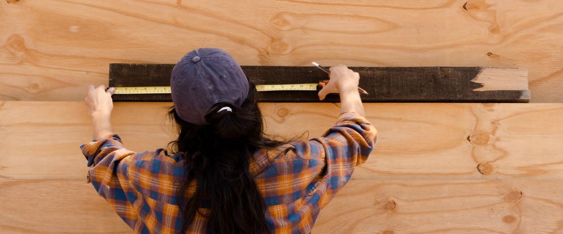 A woman seen from behind in a blue cap and an orange and blue checked shirt reaches both arms up to a dark wooden board fixed across pale plywood in daylight, holding a yellow tape measure along its length with a pencil in one hand